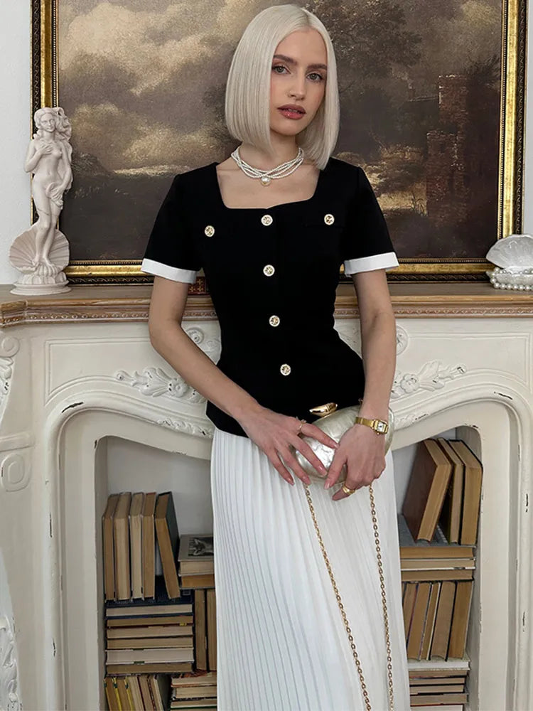 Woman in black top and white skirt standing in a room with books and a painting.