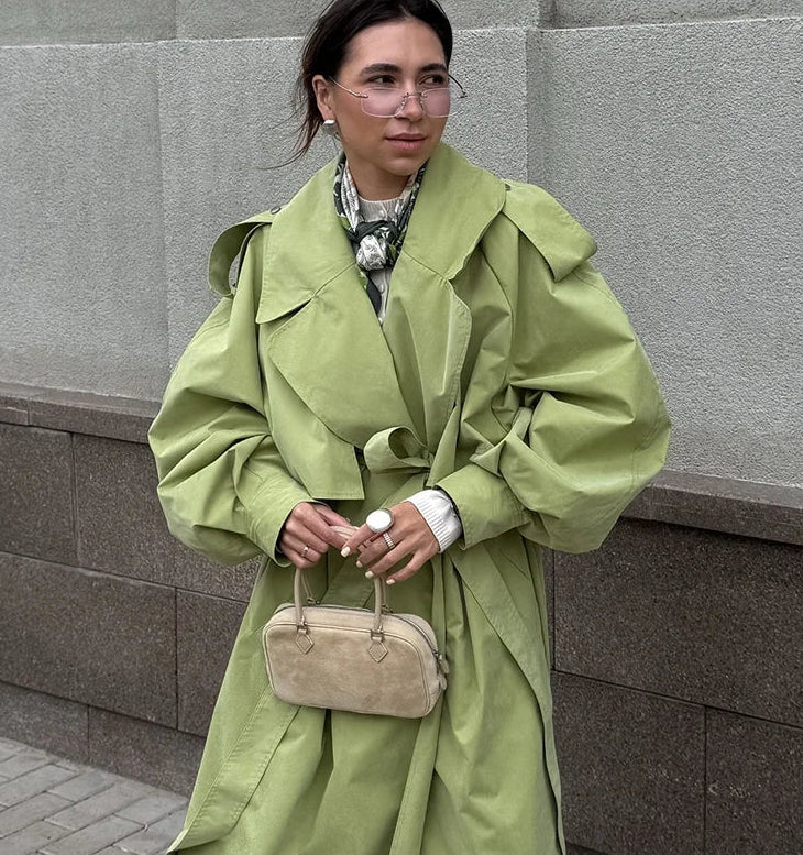 Person wearing a green trench coat holding a beige handbag against a gray wall.