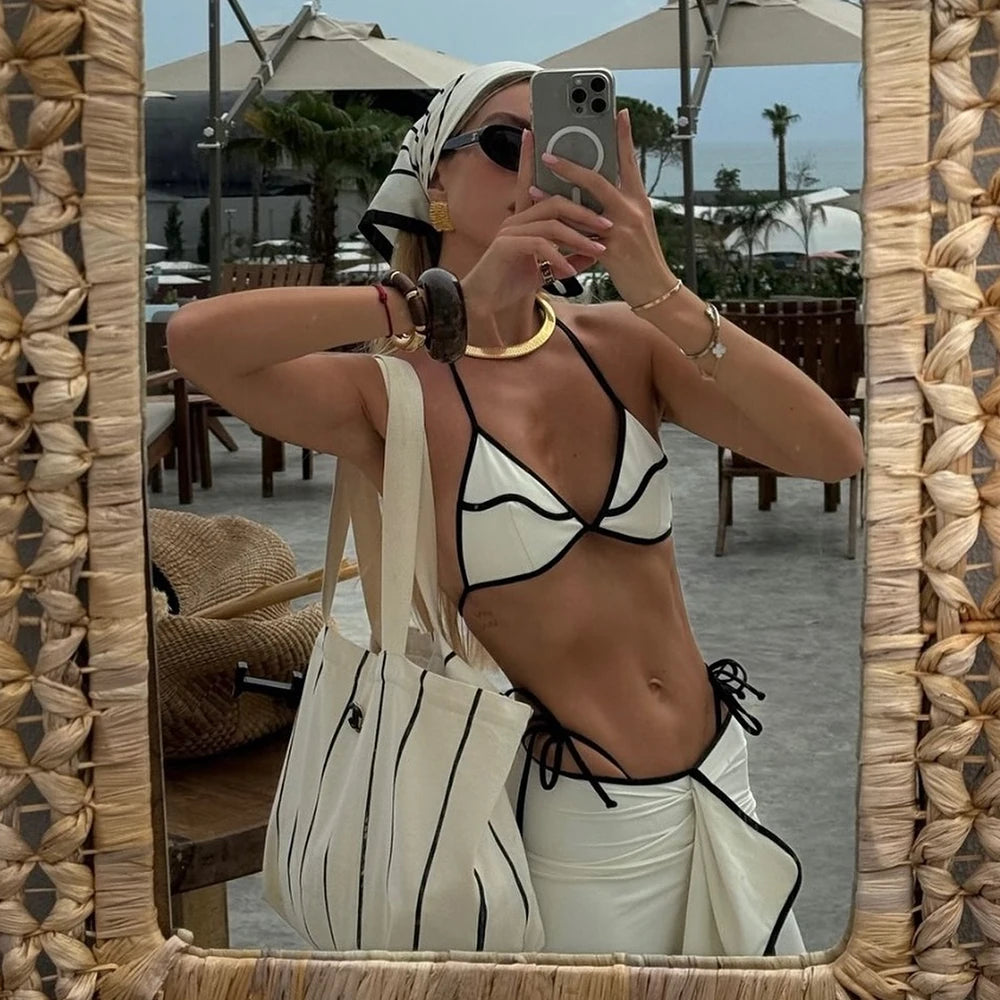 Reflection of a woman in a mirror wearing a white bikini with black contrast trim and a white ruffled maxi skirt with black trim, holding a striped tote bag.