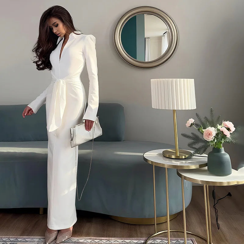 Woman in a white outfit standing in a modern living room with a lamp and flowers on a table.