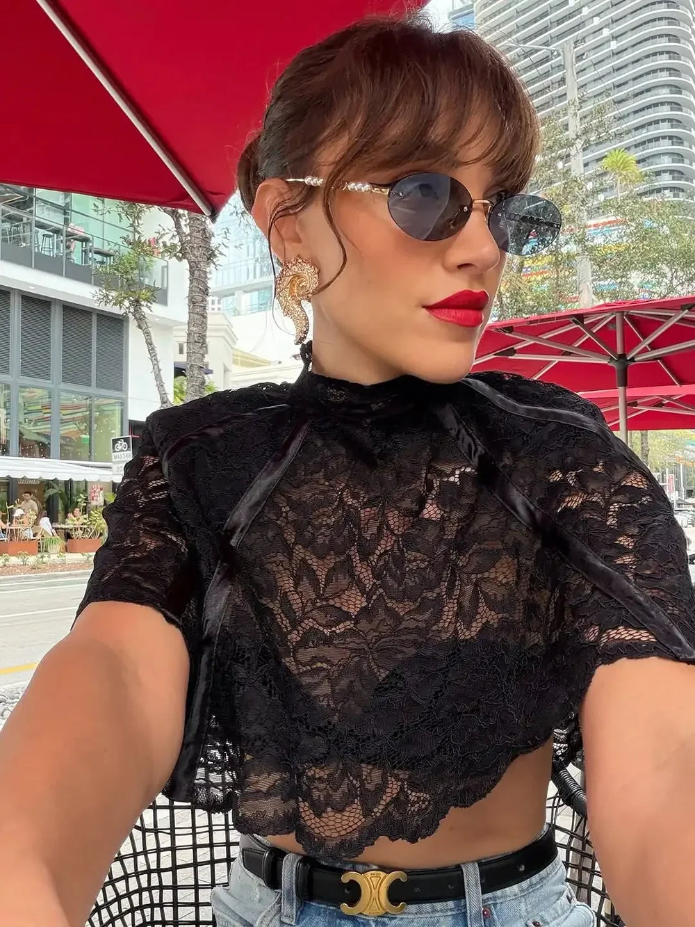 Woman wearing a black lace top with sunglasses and red lipstick, standing outdoors.