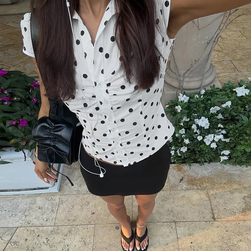 Woman wearing a white blouse with black polka dots and a black skirt, holding a black handbag.