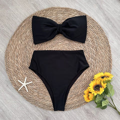 Flat lay of a black bow-front bikini top and high-waist bottoms on a woven mat.