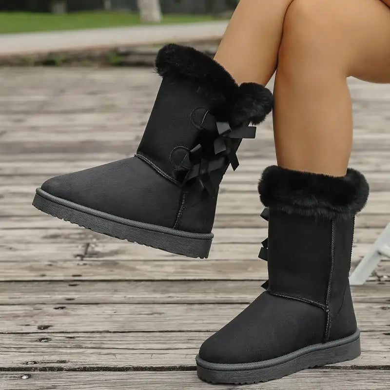 Black winter boots with fur trim worn by a person on a wooden floor.