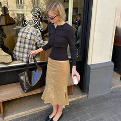 Woman in a black top and beige skirt holding a brown bag and white cup on a city street.
