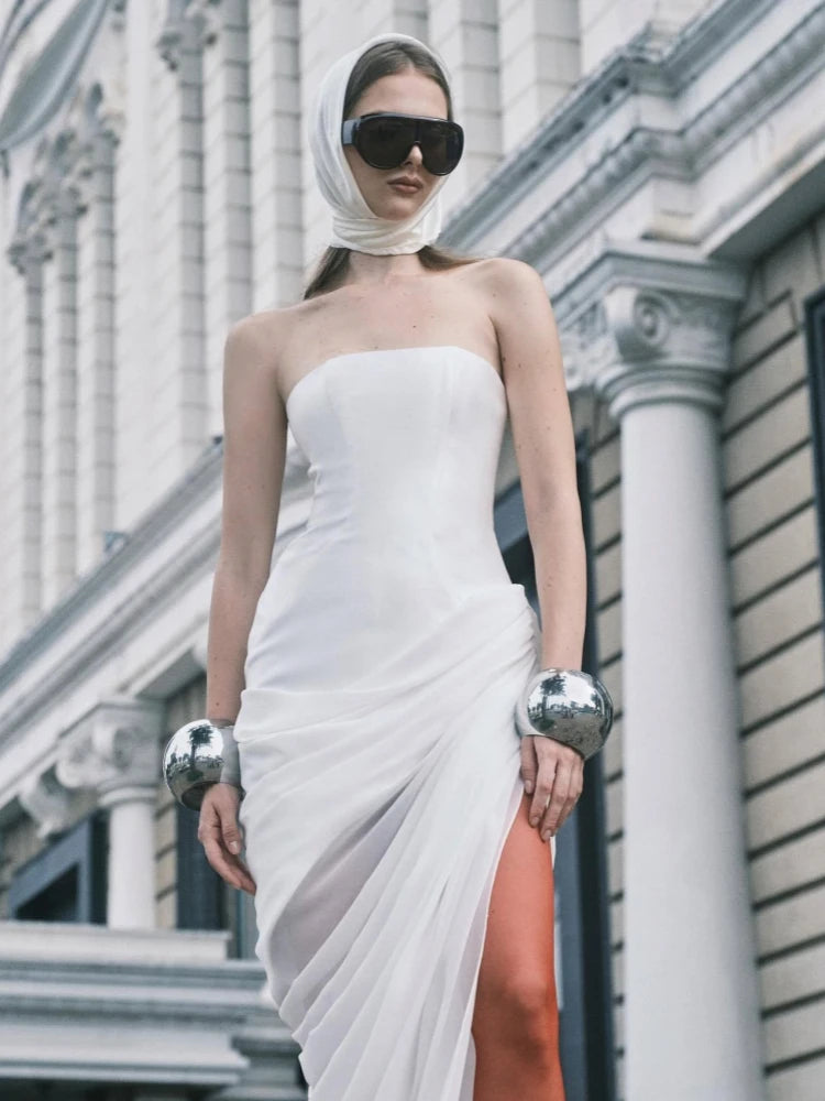 Woman in a white strapless dress with a high slit, standing in front of an architectural building.