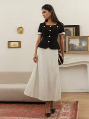 Woman in a black top and white pleated skirt standing in a room with framed pictures on the wall.