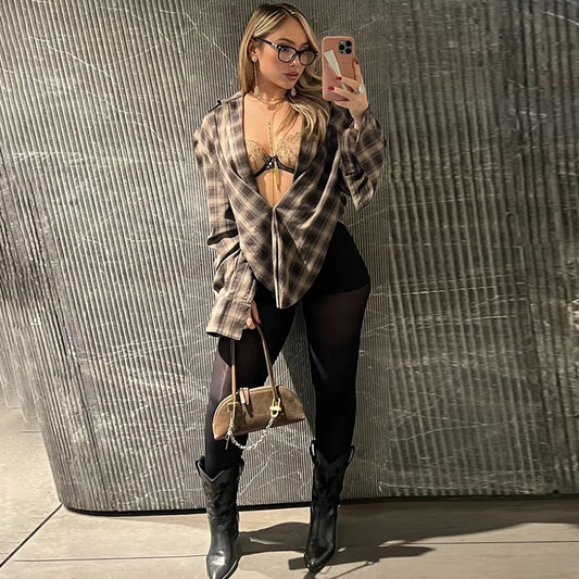 Woman taking a mirror selfie wearing a plaid shirt, black leggings, and high boots against a textured wall.