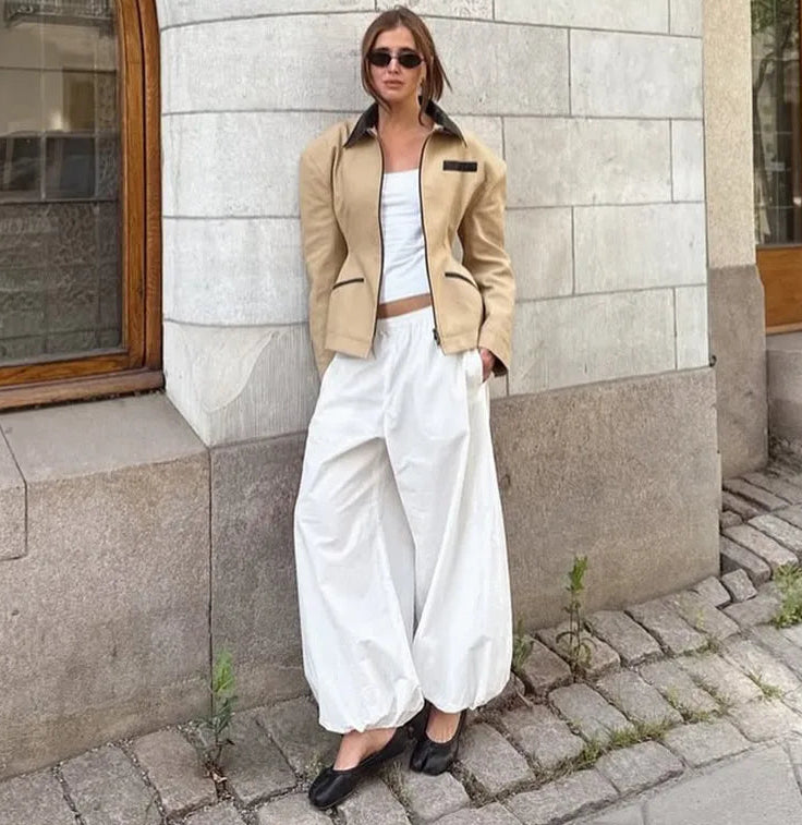 Woman in beige jacket and white pants standing against a stone wall.
