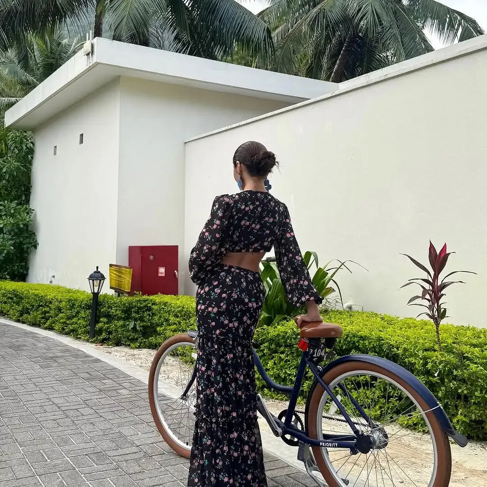 Woman in a floral dress standing next to a bicycle in front of a white wall with greenery.
