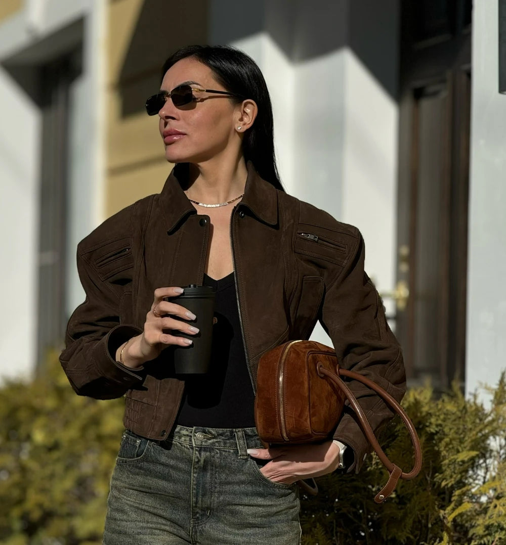 Woman in a brown jacket holding a coffee cup and a brown handbag outdoors.