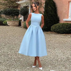 Woman in a light blue dress standing outdoors with greenery and a building in the background
