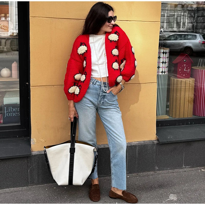 Woman wearing a red cardigan with white flowers, holding a white bag, standing against a yellow wall.
