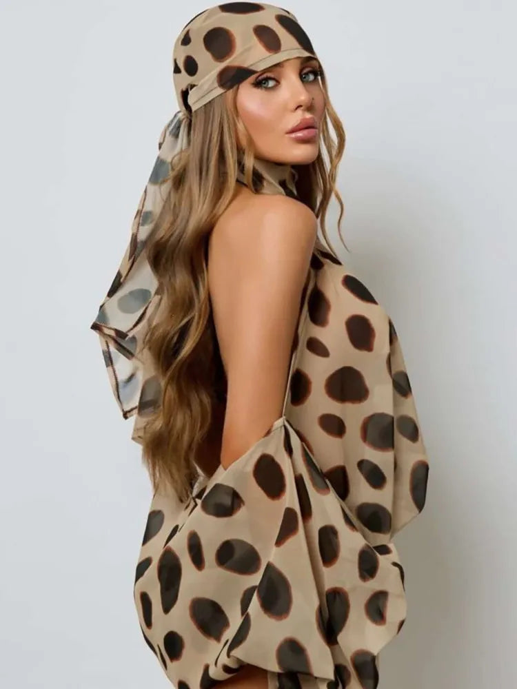 A woman wearing a beige leopard-print headscarf and matching draped wrap, photographed against a plain light gray studio background.