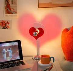 Heart-shaped lamp on a desk with a laptop and mug, surrounded by posters and a plush toy.