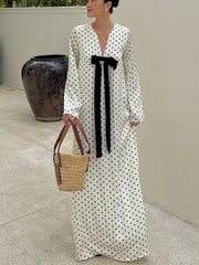 Woman wearing a long polka dot dress with a black ribbon, holding a woven bag outdoors.
