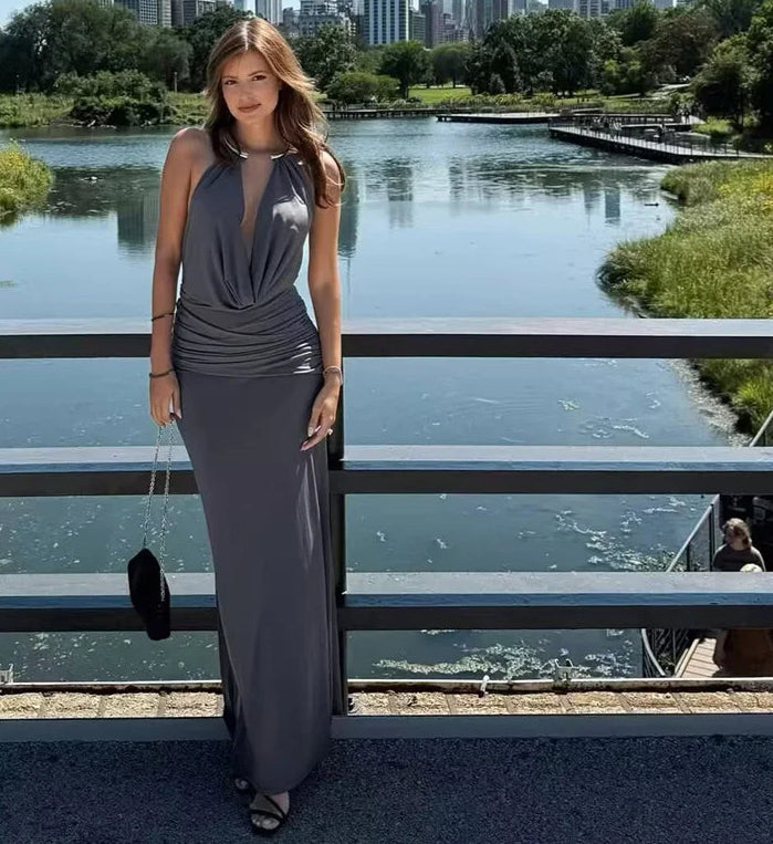 Woman in a long gray dress standing by a waterfront with greenery and buildings in the background