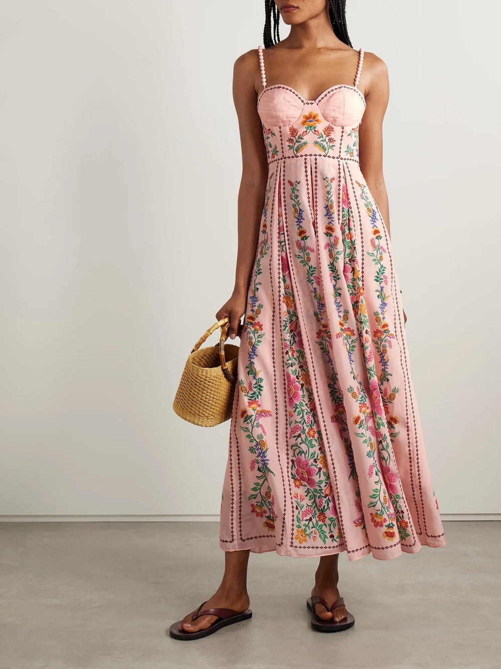 Woman wearing a pink floral dress holding a straw bag against a plain background