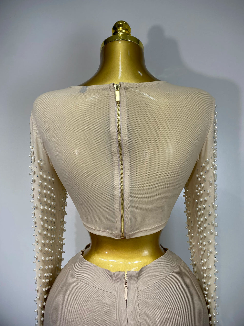 Mannequin wearing a beige outfit with pearl embellishments on sleeves against a gray background