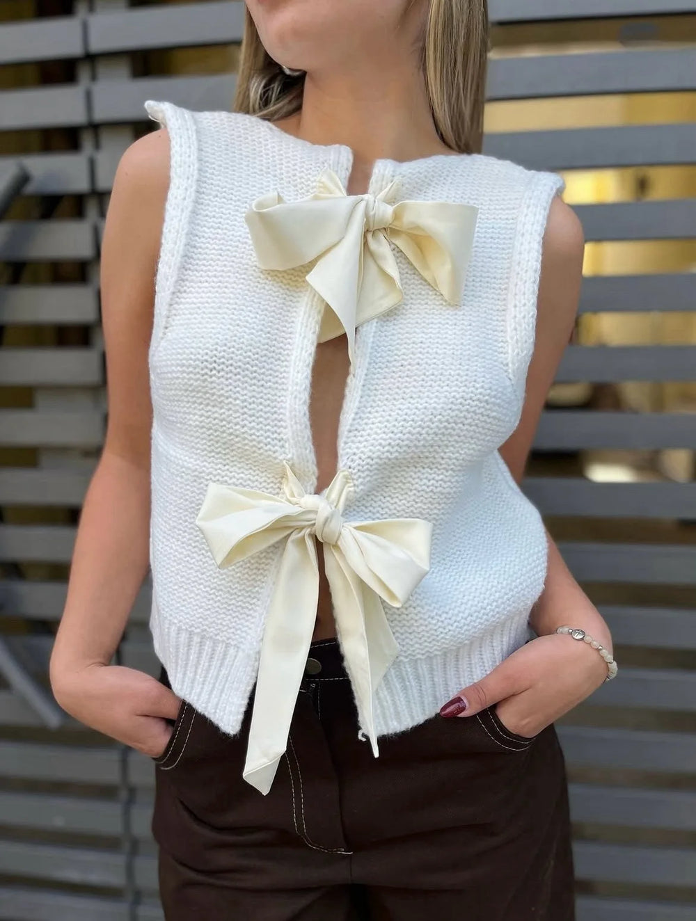 White sleeveless knitted top with bow details worn by a person, against a blurred outdoor background.