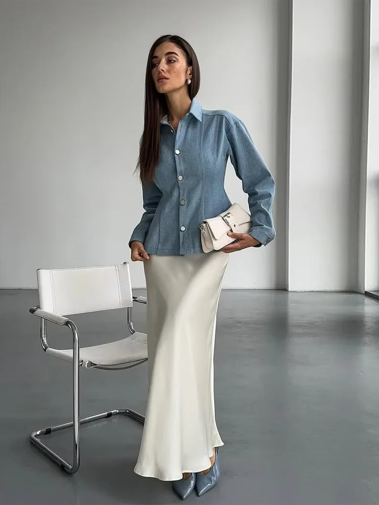 Woman in a denim shirt and white skirt holding a clutch in a minimalistic room.