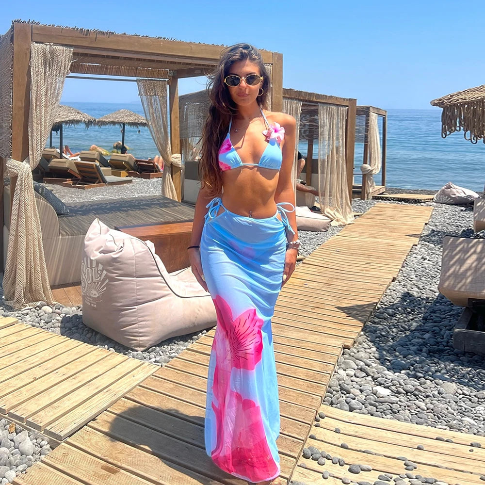 Woman in a colorful bikini and sarong standing on a wooden path by the beach.