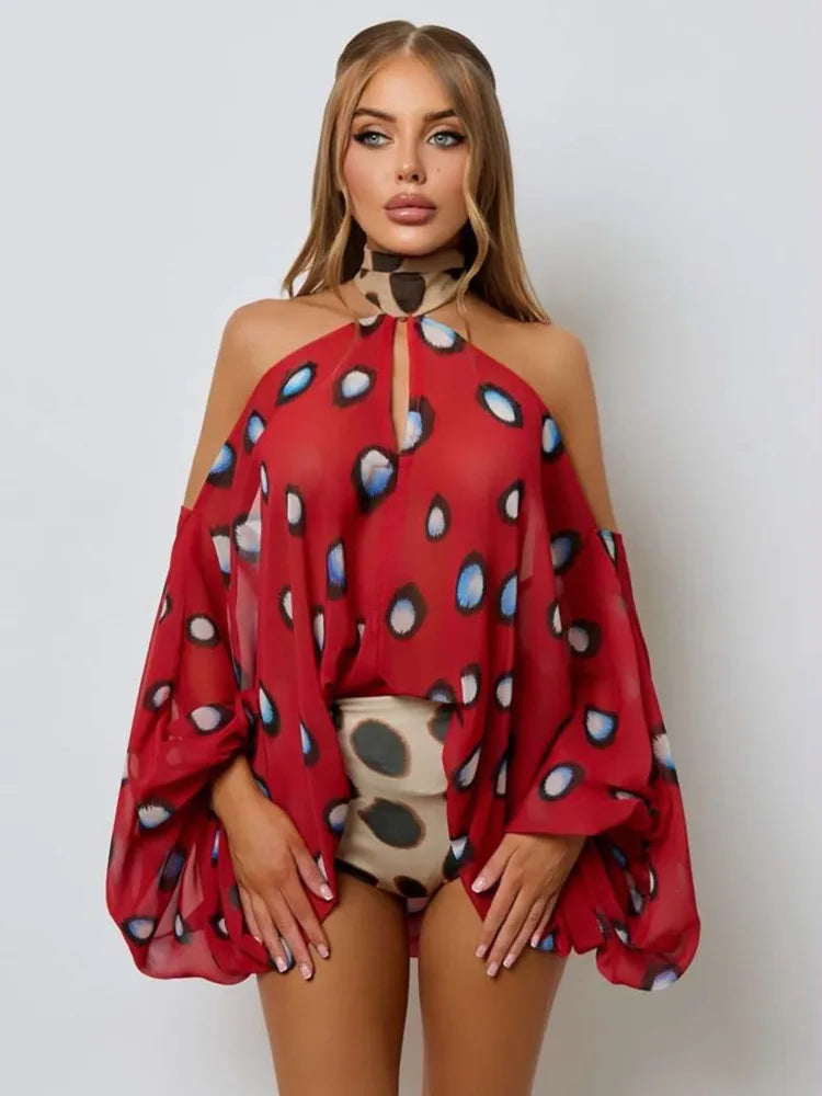 A woman modeling a sheer red cold-shoulder top with a black, white, and blue polka dot pattern, paired with a matching beige and black polka dot choker and shorts, against a light gray background.