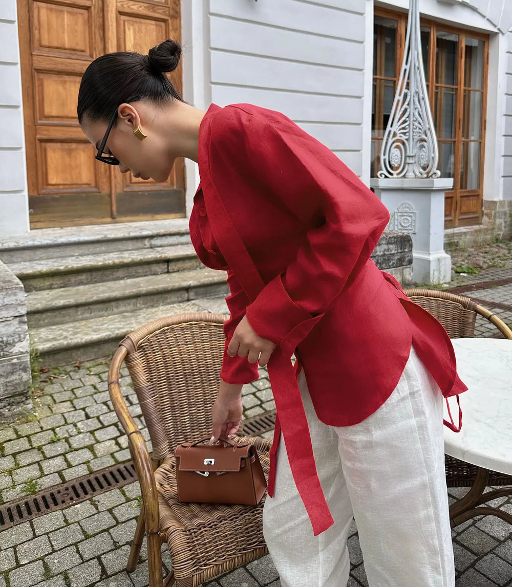Woman in a red coat and white pants standing outdoors near a wicker chair.