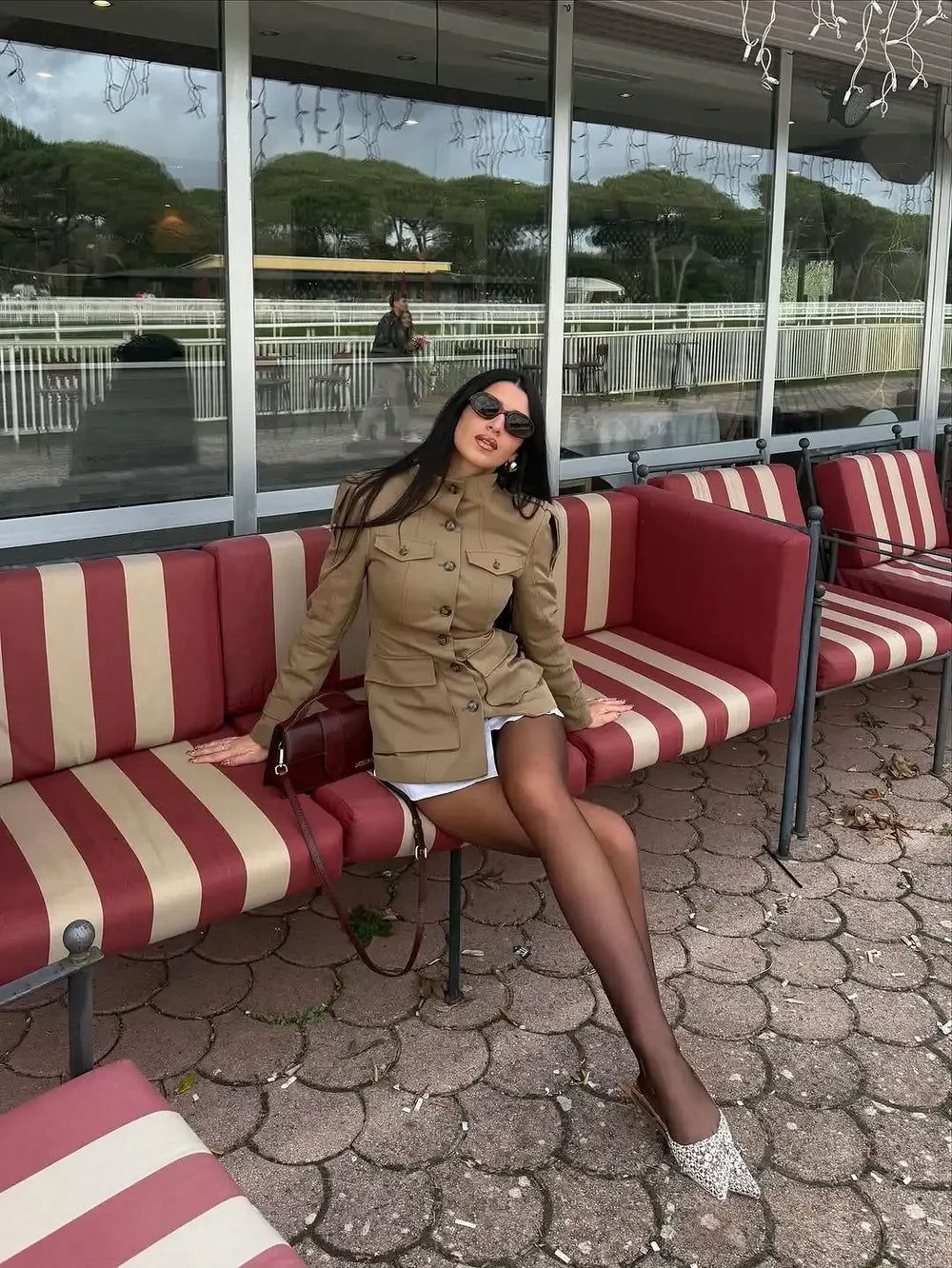 Woman sitting on a red and white striped bench in an outdoor setting with large windows in the background.