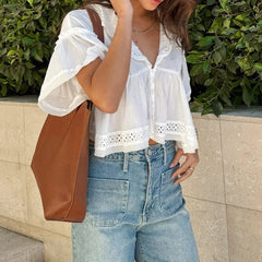 Person wearing a white blouse with lace details and blue jeans, standing against a green bush.