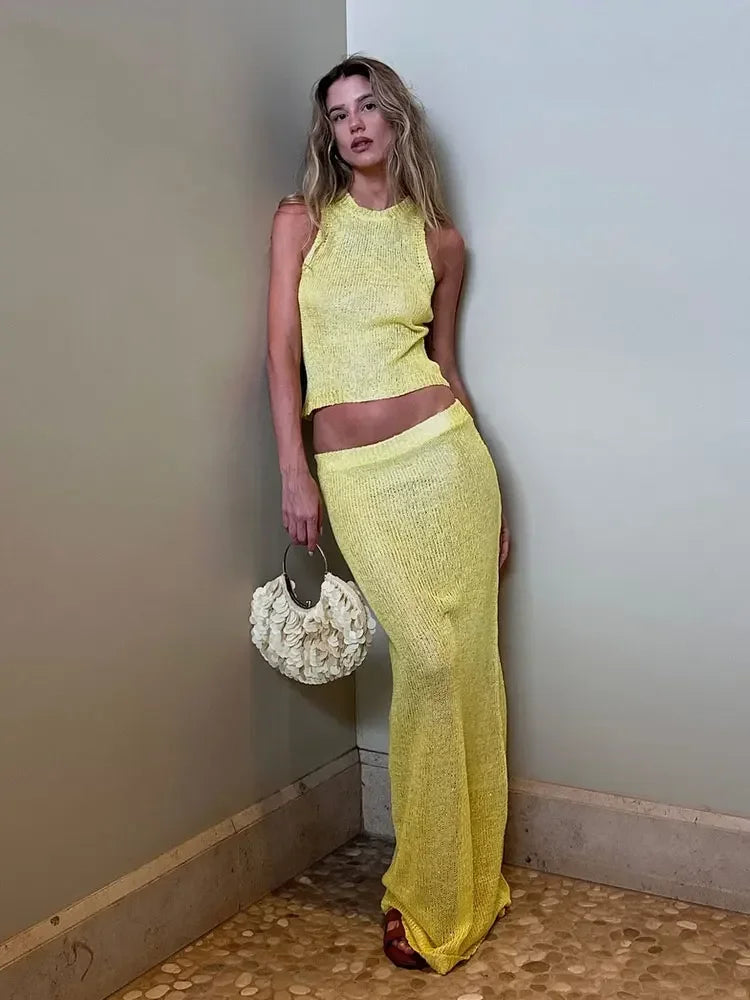 Woman in a yellow outfit holding a white handbag against a plain wall.