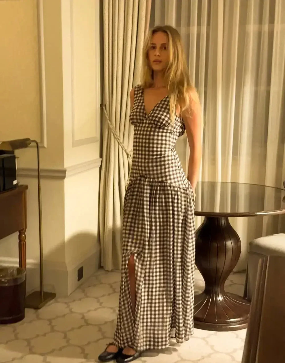 Woman in a checkered dress standing in an elegant room with curtains and a table.