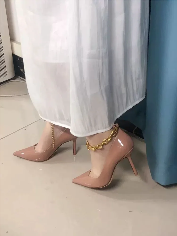 Pink high-heeled shoes with a gold anklet on a light-colored floor.