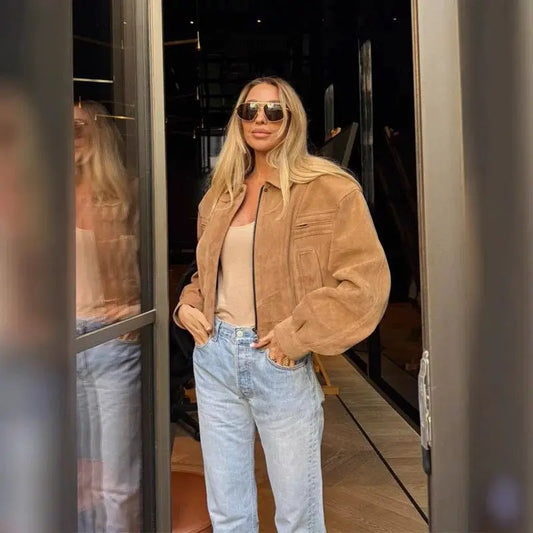 A woman wearing a khaki jacket and light blue jeans is standing in front of a mirror.