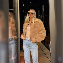 A woman wearing a khaki jacket and light blue jeans is standing in front of a mirror.