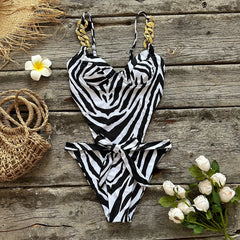 A zebra print one-piece swimsuit with gold chain straps and a tie waist, laid out on a wooden surface with beach accessories.
