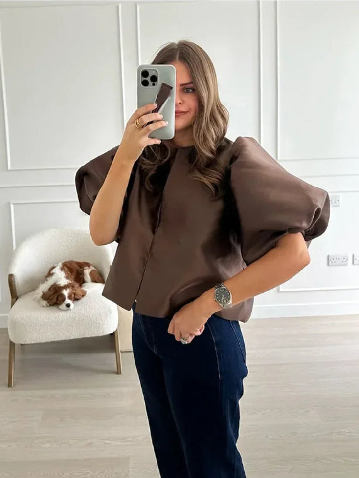 Woman taking a mirror selfie wearing a brown blouse and dark pants in a room with a dog on a chair.