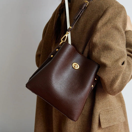 Brown leather handbag with gold accents worn over a brown coat on a neutral background