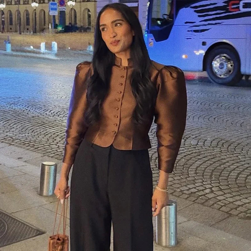 Woman in a brown blouse and black pants standing on a street with a bus in the background
