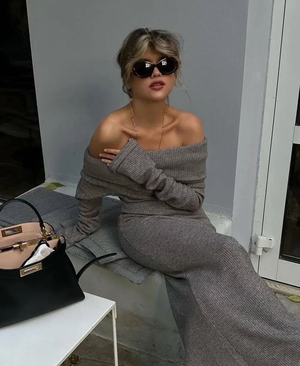 Woman in gray off-shoulder outfit sitting on a bench with a handbag next to her.