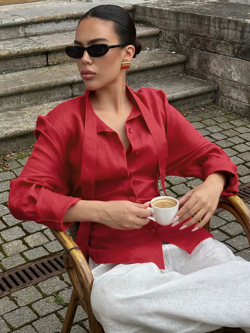 Woman in a red shirt and white pants sitting on a wooden chair holding a cup, with steps and pavement in the background.