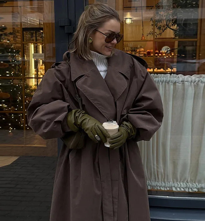 Woman in a brown coat holding a cup, standing in front of a store window.