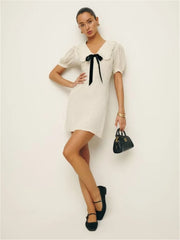 Woman wearing a white dress with a black bow, holding a black handbag on a light background