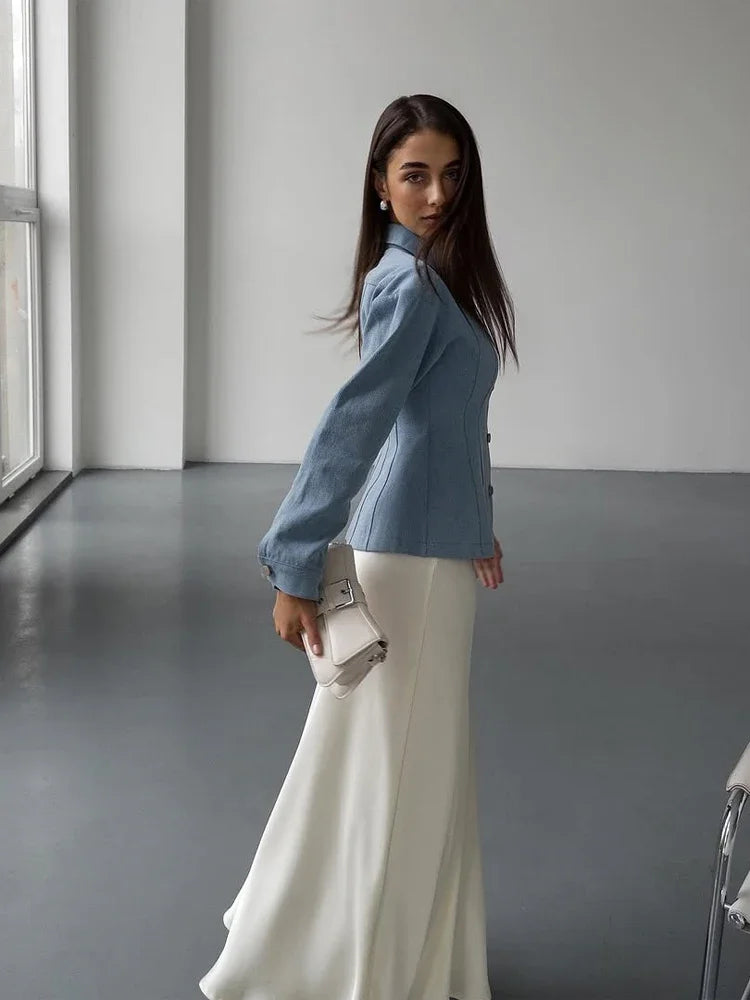 Woman in a blue blazer and white pants standing in a minimalistic room.