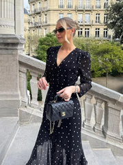Woman in a black polka dot dress holding a Chanel handbag on a balcony with classical architecture.