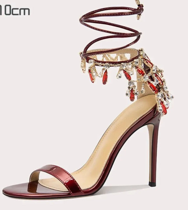 Maroon high-heeled sandal with decorative straps and red embellishments on a light gray background.