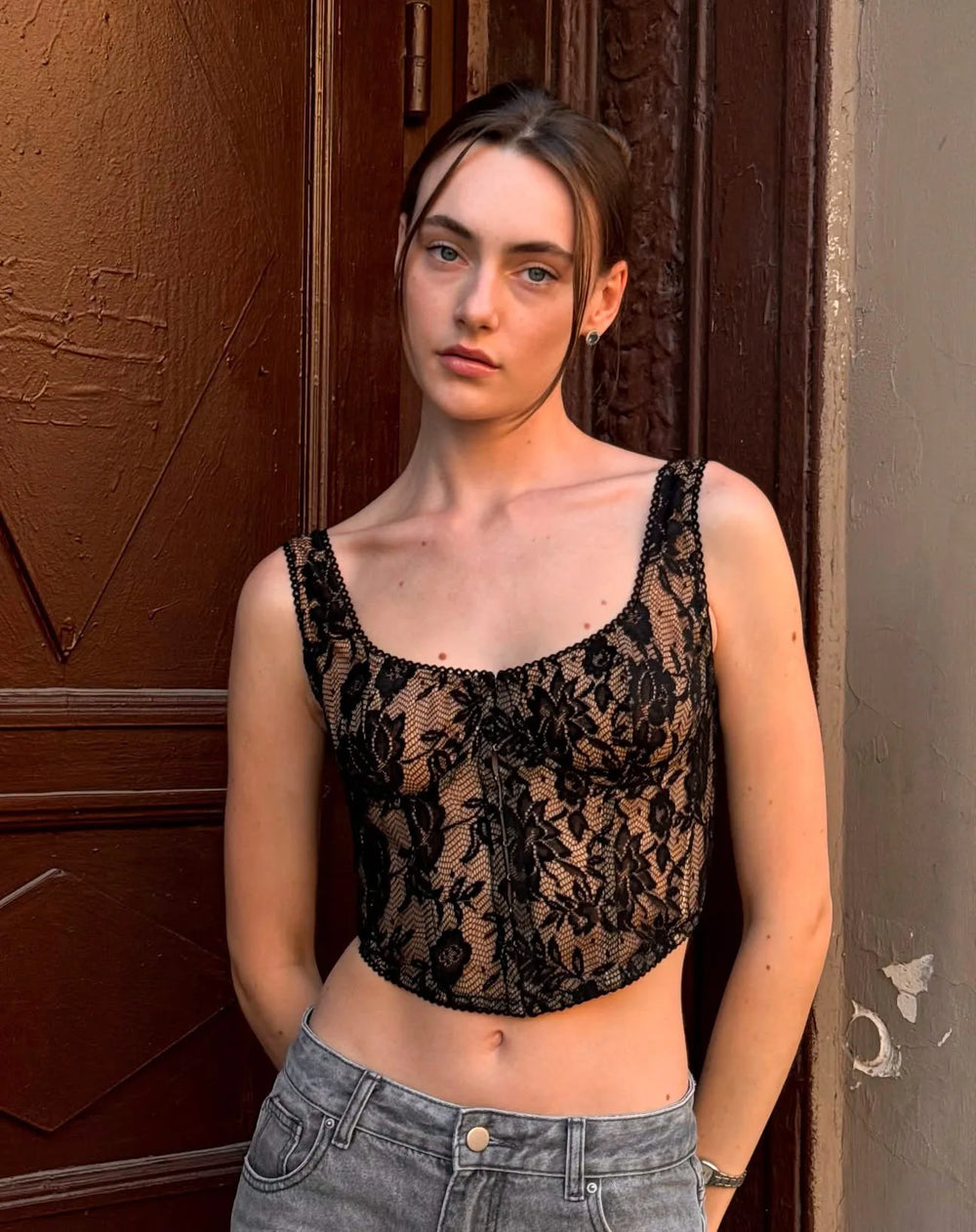 Woman wearing a black lace top and gray jeans standing against a textured wall.