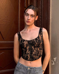 Woman wearing a black lace top and gray jeans standing against a textured wall.