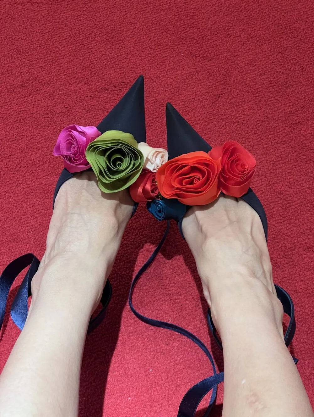Close-up of dark blue stiletto heels with colorful fabric roses and ankle straps on a red carpet.