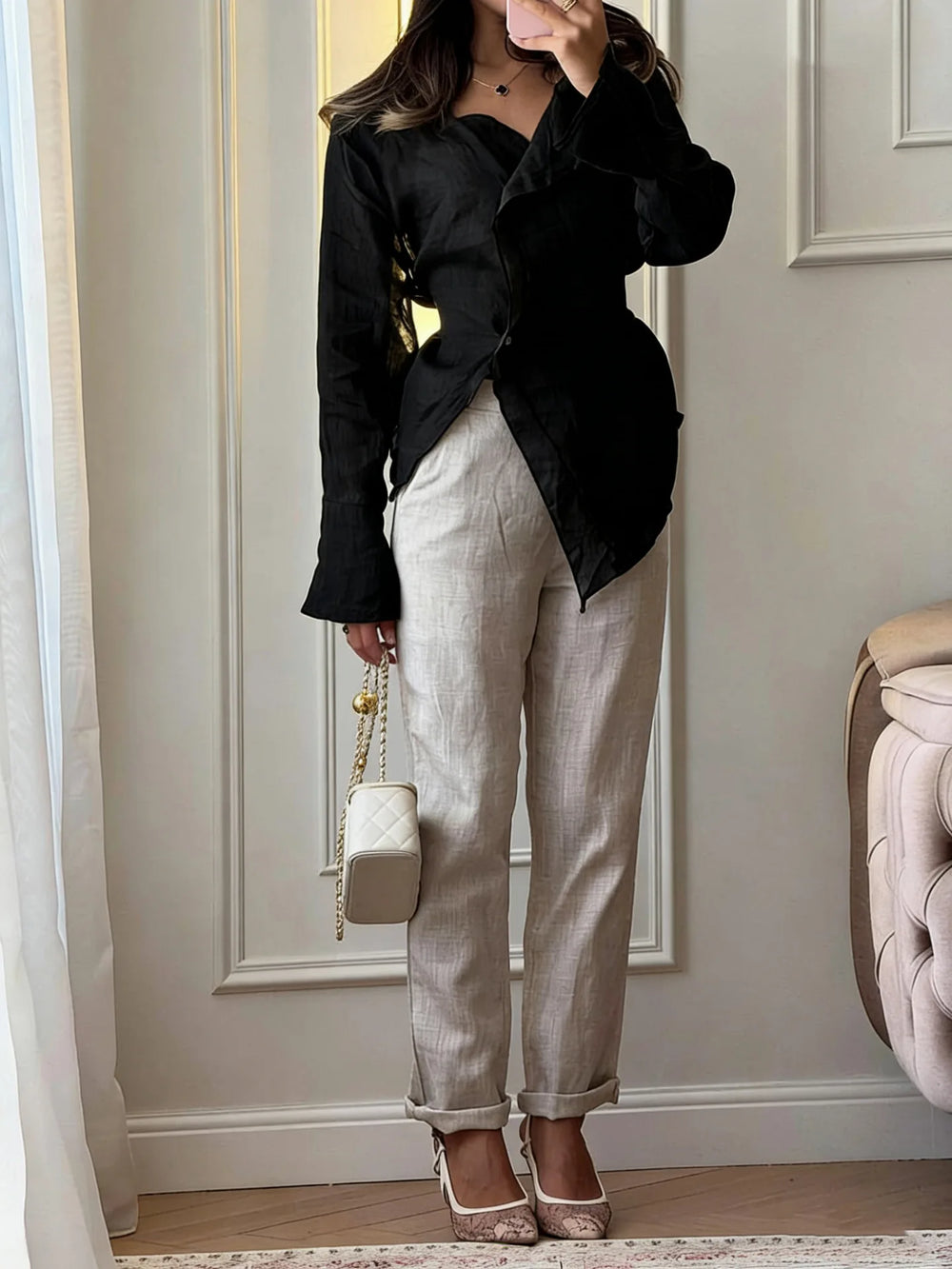 Person wearing a black top and beige pants, holding a white handbag, standing in a room with light-colored walls and a chair.